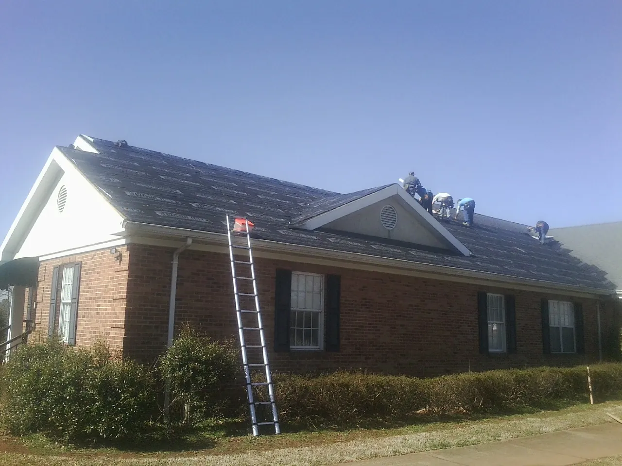 Crew tearing shingles off a brick ranch roof with a ladder set — Metal Roof Repair work by our crew in Avon